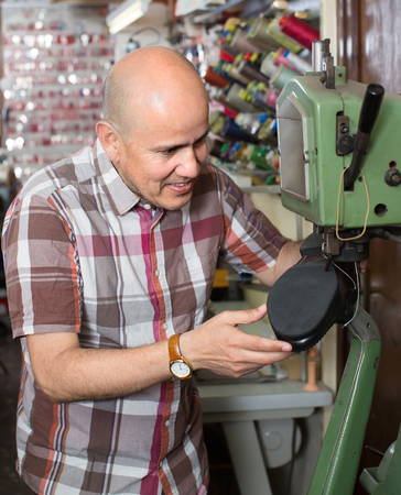 Smiling mature repairman stitching footwear on machine in shoe atelierの写真素材