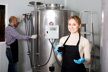 Young pretty woman wearing uniform standing among brewery stainless equipmentの写真素材