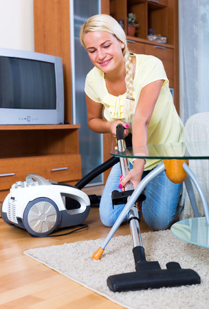 Cheerful blonde young woman hoovering in living room and smilingの写真素材