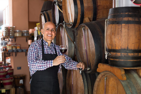 Friendly smiling mature  seller tasting wine from wood barrel  in wine houseの写真素材