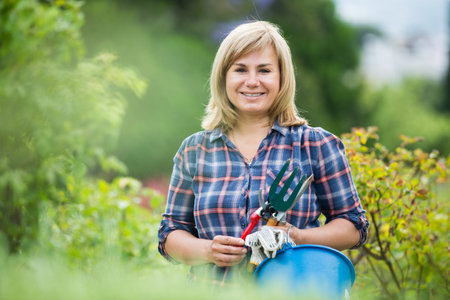 glad blond mature woman having horticultural instruments in garden on summer dayの写真素材