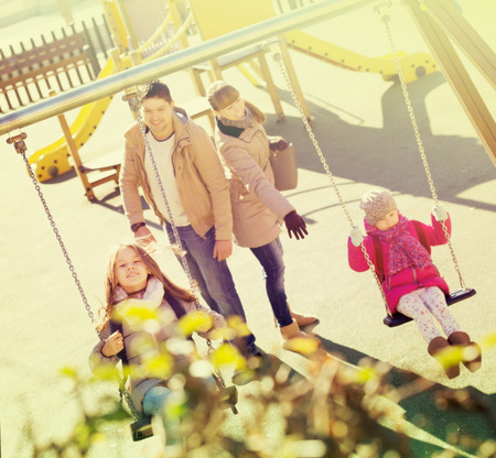 Positive young parents watching little daughters swinging at playgroundの写真素材