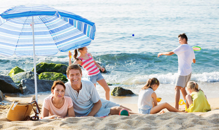 Happy couple relaxing on beach while their four kids playing active games and sand on summer dayの写真素材
