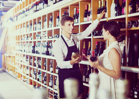 Smiling young seller man wearing apron helping to buy bottle of wine to woman customer in wine storeの写真素材