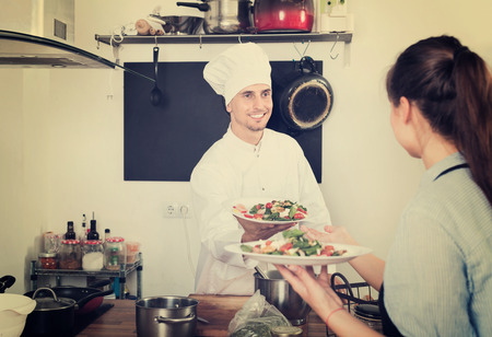 Portrait of glad young smiling man cook giving to waitress ready to serve saladの写真素材