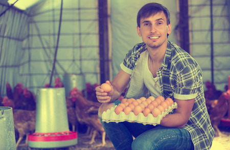 Young man farmer holding container with fresh eggs in hands in henhouseの写真素材