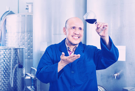 Smiling mature man holding glass of red wine and standing in winery production section