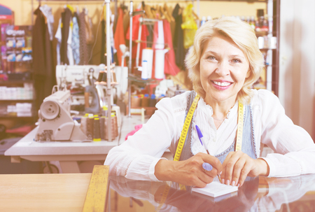 Smiling mature woman tailor taking order at counter in sewing workshopの写真素材