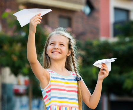smiling girl in bright dress playing with flying paper airplane toy outdoorsの写真素材