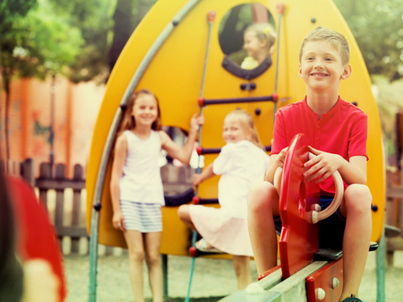 portrait of glad boy sitting on swinging toy while other children playing outdoorsの写真素材