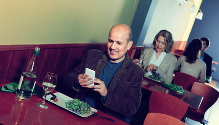 Smiling mature man taking pictures of dinner with smartphone in restaurant