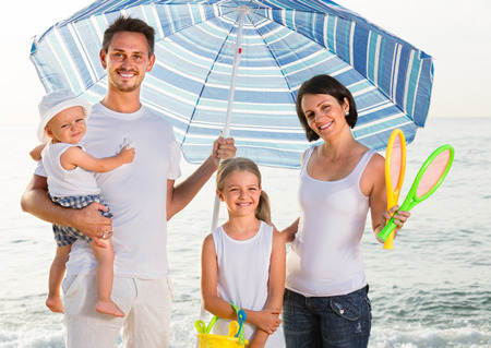 positive family of four standing with plastic bucket and scoops under sun umbrella at beachの写真素材