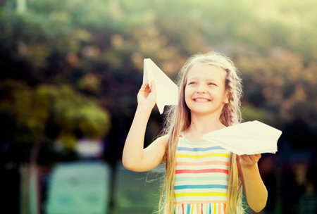 smiling girl in bright dress playing with flying paper airplane toy outdoorsの写真素材
