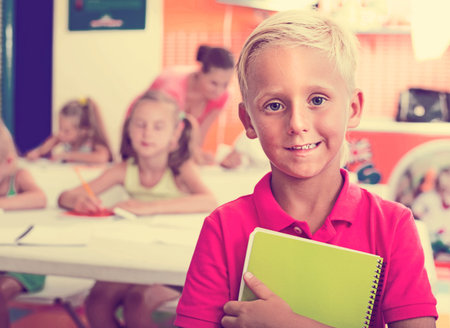 Smiling boy pupil standing in elementary school class with book in hands