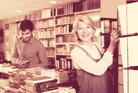 mature woman standing among bookshelves and looking for book