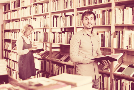 Young  cheerful positive man reading book while choosing it in book shopの写真素材