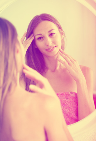 young positive russian woman examining her face by looking at it in mirrorの写真素材