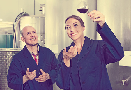 Two cheerful winery workers holding glass of wine in fermenting section of factory. Focus on womanの写真素材