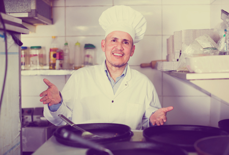smiling mature man chef in uniform standing with frying pans in restaurant's kitchenの写真素材