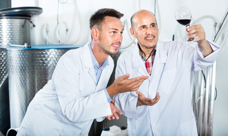 Happy two factory workers in the uniform standing together and examining sample of wine in glass. Focus on young manの写真素材