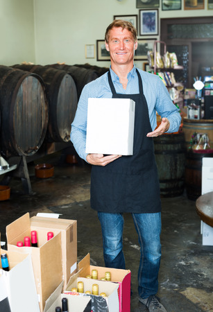 Smiling seller man in apron carrying carton package with bottles in wine houseの写真素材