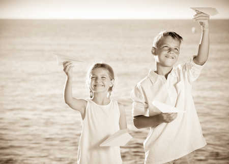 portrait of smiling boy and girl holding paper plane toys in hands on beachの写真素材