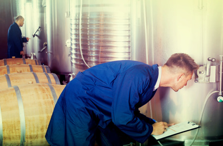 Portrait of adult man expert wearing coat standing and taking off data from equipment in wineryの写真素材