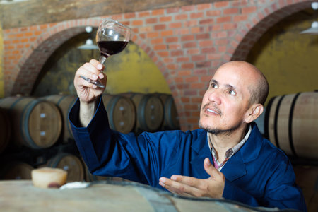 smiling mature winemaker in working coat looking at glass of wine in cellar