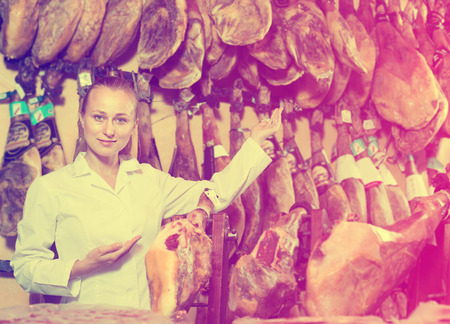 Cheerful woman seller wearing uniform showing iberian ham in meat shopの写真素材