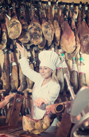 Positive young woman wearing uniform showing iberian ham in meat shopの写真素材