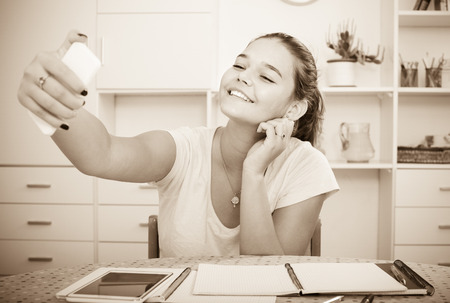 cheerful smiling female teenager doing selfie during homework at the tableの写真素材