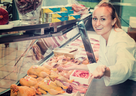 cheerful young woman wearing coat selling fresh chicken carcass in delicatessen sectionの写真素材