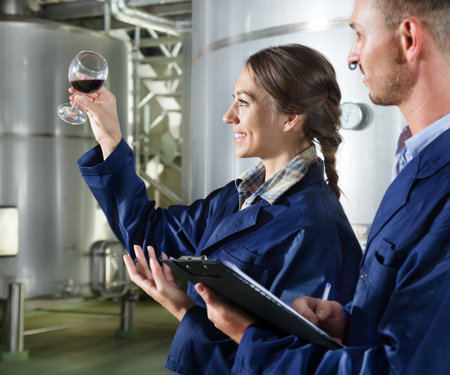 Young man and woman employees looking at wine sample in the glass and taking notes on a winery...