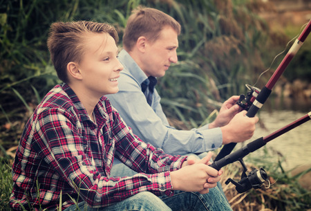 Glad father and his teenager son fishing together from water side on a river. Focus on childの写真素材