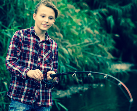 Portrait of positive teenage boy fishing on freshwater lake from shoreの写真素材