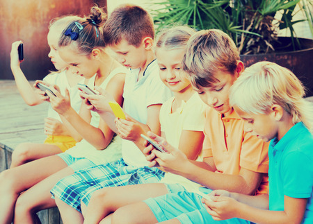 Busy cute children holding smartphones in hands and sitting together outdoorsの写真素材