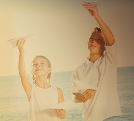 happy two kids in elementary school playing together with origami airplane toys on beachの写真素材