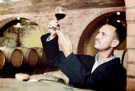 Adult male winemaker in uniform having glass of wine in hands in cellarの写真素材