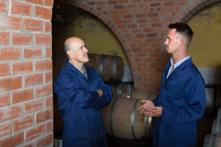 two smiling men professionals in uniforms standing in cellar with wine woods