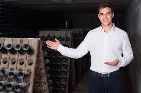 Young cheerful smiling man wine maker standing in cellar with wine bottles storage