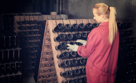 Happy woman winery employee in coat working in aging section with bottle racks in cellarの写真素材