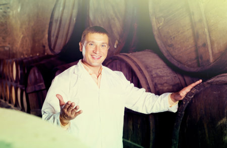 Smiling worker of winery posing with wine barrels in cellar