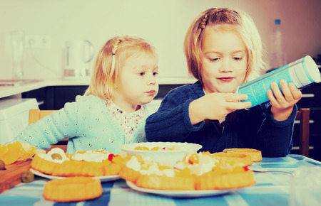 Laughing small sisters enjoying pastry with cream in kitchenの写真素材