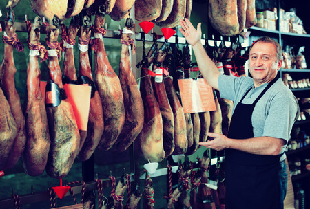 Portrait of cheerful  smiling mature shop man offering Spanish jamon in deliの写真素材