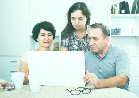 Positive elderly man and woman looking at laptop together with their adult daughter indoors. Focus on girlの写真素材
