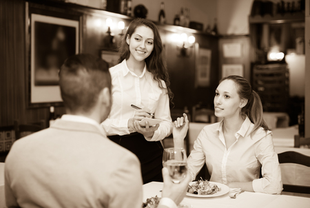Cheerful waitress offering to young couple tasty dishes. Focus on girlの写真素材