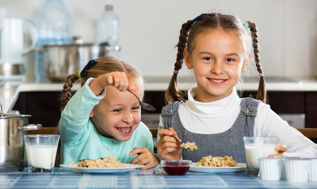 Two small cheerful girls eating healthy oatmeal at home kitchenの写真素材