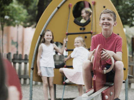 portrait of glad boy sitting on swinging toy while other children playing outdoorsの写真素材