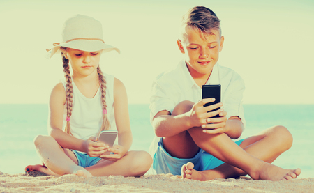 cheerful  boy and girl playing with mobile phones on sandy beachの写真素材