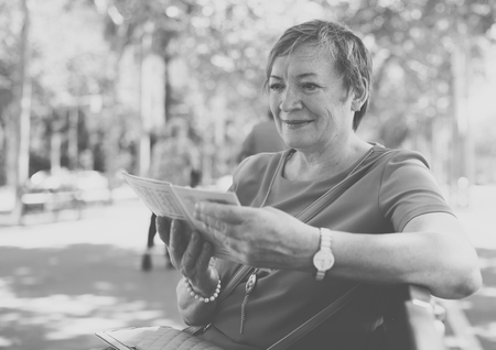 portrait of cheerful senior woman sitting on bench and reading outdoorsの写真素材